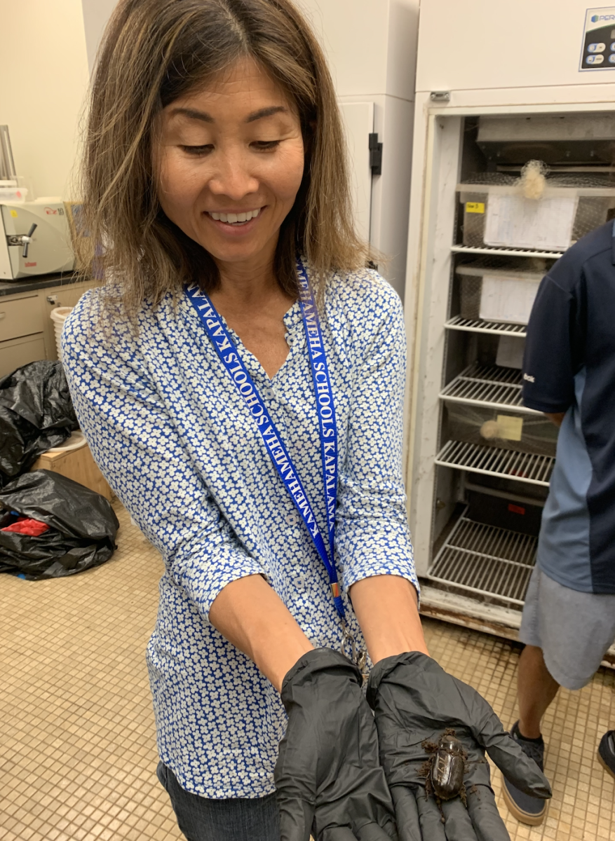 “Sally Nhomi, internship coordinator, holding a coconut rhinocerous beetle (CRB), (an invasive species threatening coconuts, statewide) during visit to the UH Mānoa College of Tropical Agriculture and Human Resources (CTAHR) department in Spring ʻ23.  The visit included Jade Hao, KS ʻ23, who served as a CTAHR intern who created a video on the importance of niu for Hawaiians, with a kāhea to halt the spread of CRB.”